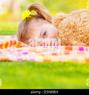 Pretty small girl lying down on green field, playing game with big brown teddy bear, cute child enjoying spring nature