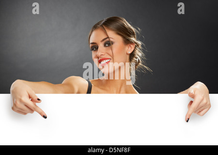 Excited young woman pointing her fingers down to the blank white advertising banner sign against dark background. Stock Photo