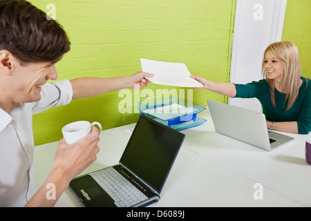 Happy young businesspeople passing documents each other with laptops t desk Stock Photo