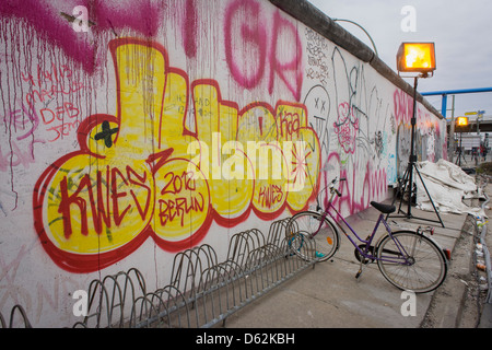 Where young Germans once risked their lives, graffiti and tags now adorn the concrete surfaces of original sections of the Berlin wall at the East Side Gallery on Muhlenstrasse, Berlin. The site is the former border between Communist East and West Berlin during the Cold War. The Berlin Wall was a barrier constructed by the German Democratic Republic (GDR, East Germany) starting on 13 August 1961, that completely cut off (by land) West Berlin from surrounding East Germany and from East Berlin. . (More in Description). . Stock Photo