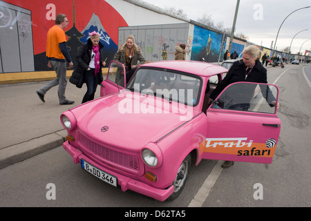 Visitors enjoy the art and an old Trabant car at the old Berlin Wall at the East Side Gallery, the former border between Communist East and West Berlin during the Cold War. Trabants were the common Socialist vehicle in East Germany, exported to countries both inside and outside the communist bloc. The Berlin Wall was a barrier constructed by the German Democratic Republic (GDR, East Germany) that completely cut off (by land) West Berlin from surrounding East Germany and from East Berlin .. (More in Description). Stock Photo