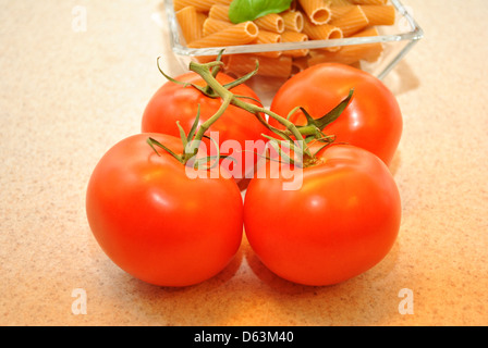 Four Plump Tomatoes in Front of a Bowl of Wheat Pasta Stock Photo