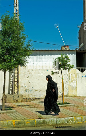 An old woman in a traditional black clothes walks down the street in the center of Erbil, Iraq along side a wall marked bullets. Stock Photo