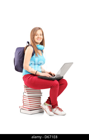 A smiling student with backpack working on a laptop and sitting on pile of books, isolated on white background Stock Photo