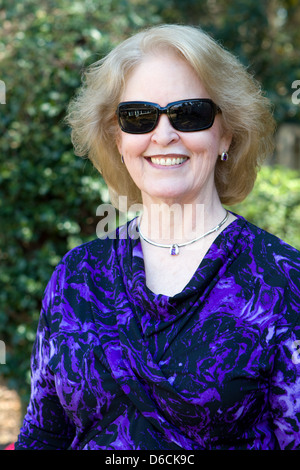 Senior woman wearing sunglasses stands posing in the backyard outside her home and smiles. Stock Photo
