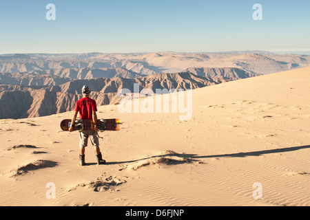 Young man holding a sandboard looking at the view from Cerro Blanco, a sand dune on the top of a mountain with 2078 meters high. Stock Photo