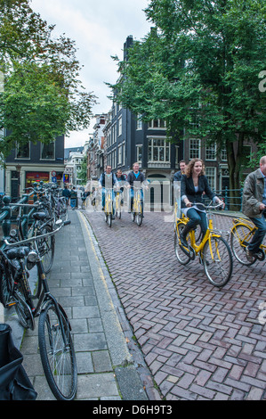 Cyclists riding over a canal bridge in Amsterdam, bicycles chained to side railings along the way. Stock Photo