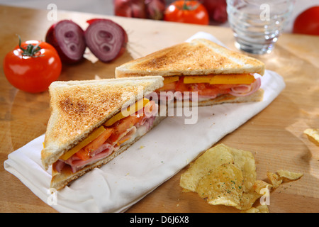 Toasted  ham cheese tomato sandwich on a white napkin on a wooden breadboard with whole tomatoes and halved red onions and crisp Stock Photo