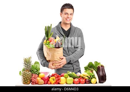 Handsome young man with a bag of groceries standing behind a pile of fruits and vegetables, isolated on white background Stock Photo