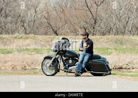 A man in his late 20s or early 30s, sits sideways on his Harley Davidson motorcycle and enjoys the early spring day. Oklahoma, USA. Stock Photo