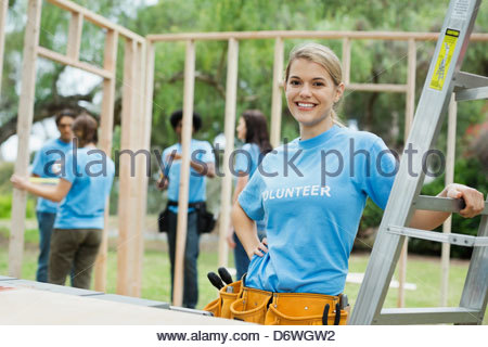 Portrait of confident woman by ladder with volunteers building wooden frame in background