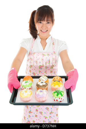 Young Asian female baking bread and cupcakes, wearing apron and gloves holding tray isolated on white. Stock Photo