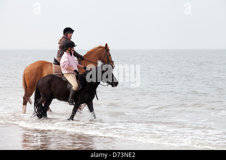 Two young women riding horses in the sea surf at the waters edge, Holkham Beach, Norfolk UK Stock Photo