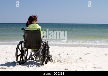 Disabled woman sits alone in a wheelchair on a sandy beach watching the ocean waves. Stock Photo