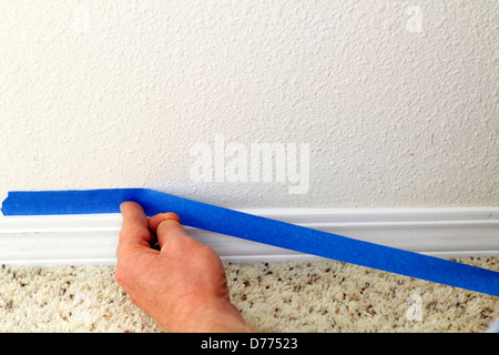 Taping painters tape to wall above baseboard. Male hand preparing to paint wall trim by using blue painter's tape on wall above it for protection. Stock Photo