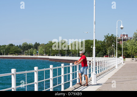 A man stands looking out over the railing at the beautiful blue harbor on the boardwalk in Sarnia Ontario, Canada Stock Photo