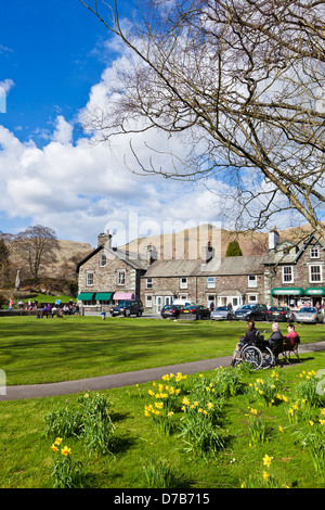 People sat on benches around the village green enjoying the spring sunshine  in Grasmere Cumbria England UK GB EU Europe Stock Photo