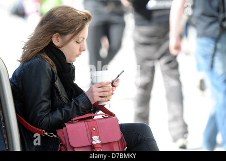 A girl  sits on a bench with a coffee in her hand while using a phone. Stock Photo