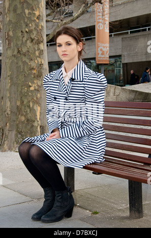 A young woman with a serious expression and a striped coat sits on a bench on the south Bank in London Stock Photo