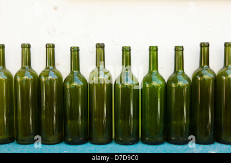Ten empty green wine bottles in a row on the background of old run-down white wall, resting on turquoise wooden surface. Stock Photo