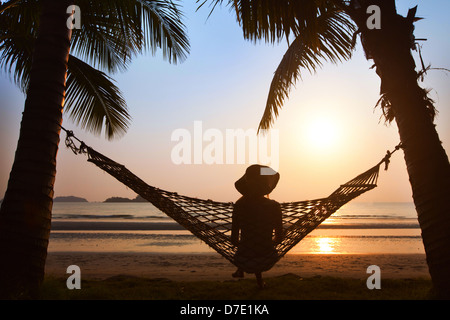 silhouette of woman in hat sitting in hammock at sunset on the beach Stock Photo