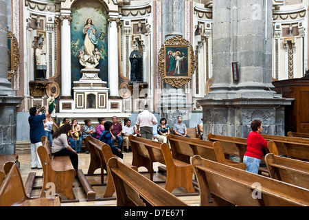 diverse Mexican tour group sit in side aisle as guide lectures & parishioners pray  Church of the Brotherhood of Jesus Puebla Stock Photo