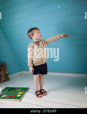 A little boy in nice clothes and sandals is playing alone with old fashioned board game in a blue and white loft room. Stock Photo