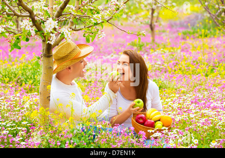 Two lovers on picnic in beautiful spring garden, purple flowers field, blooming tree, handsome man feeding girlfriend apple Stock Photo