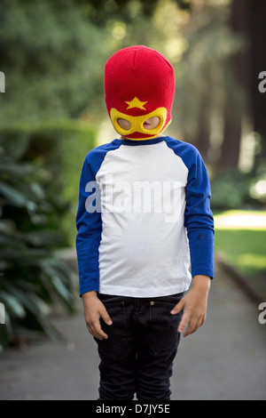 little boy wearing red and yellow superhero mask posing outside on the sidewalk, obscuring his eyes Stock Photo