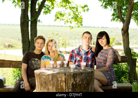 Group of young friends enjoy a drink while seated outdoors on a rustic wooden bench around a tree stump table Stock Photo
