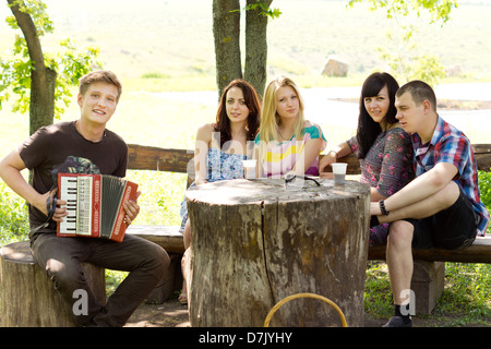 Young man sitting under the shade of a tree playing the accordion for a group of his friends as they enjoy a day out in the country. Stock Photo