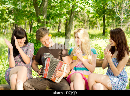 Three beautiful young female friends enjoying music played on a concertina accordion by a handsome young man as they clap to the rythm. Stock Photo