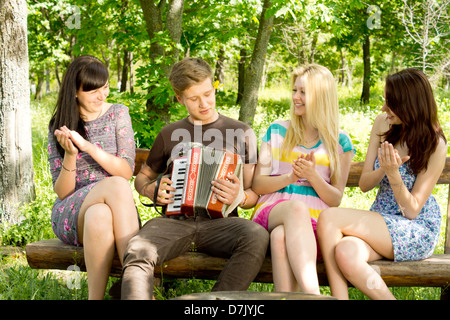 Happy young woman friends clapping along to the music played on an accordion by a handsome young man as they enjoy a relaxing day in the park. Stock Photo