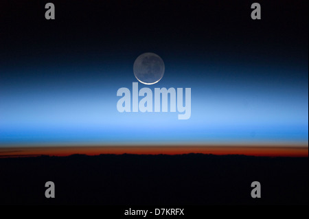 Moon with the limb of Earth near the bottom transitioning into the orange-colored troposphere, the lowest and most dense portion of the Earth's atmosphere. The troposphere ends abruptly at the tropopause, which appears in the image as the sharp boundary between the orange- and blue- colored atmosphere. The silvery-blue noctilucent clouds extend far above the Earth's troposphere. Stock Photo