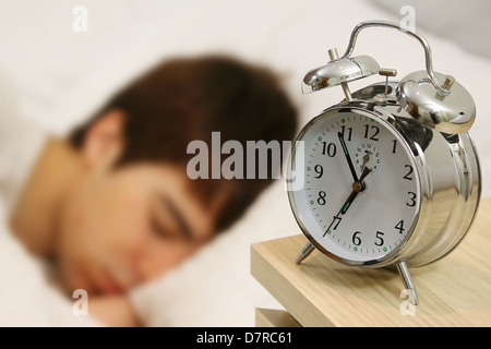 Young male adult asleep in bed, an alarm clock by his bed just before 7am. The alarm is just about to ring. Stock Photo