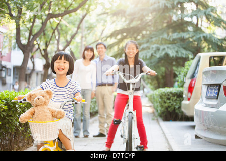 Sisters Ride Their Bicycles While Parents Watch Stock Photo