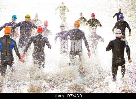 Triathletes in wetsuits running into ocean Stock Photo