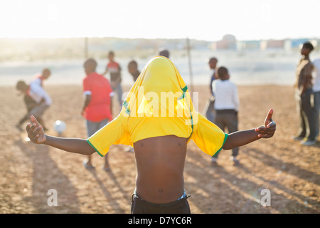 Boy celebrating with soccer jersey on his head in dirt field Stock Photo