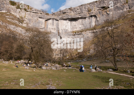 Sunny view of people visiting & relaxing at scenic Malham Cove - huge, sheer, curving limestone cliff in beautiful Yorkshire Dales, England, GB, UK. Stock Photo