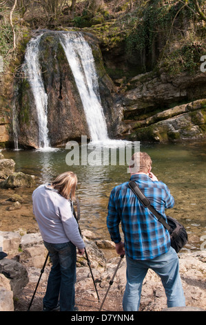 Rear view of 2 photographers using tripods (man & woman) taking photos of scenic waterfall - Janet's Foss, Malham, Yorkshire Dales, England, UK.