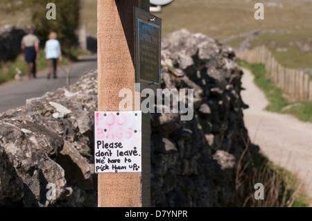 Close-up of handwritten note attached to wooden post for dog owners to remove poo dirt bags + dog walkers in background - Malham, Yorkshire Dales, UK Stock Photo