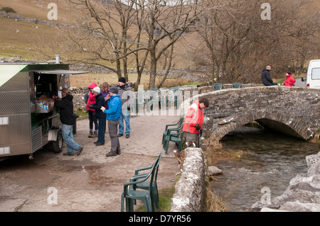 Walkers buying refreshments from mobile snack-bar by beck flowing under old rustic stone Gordale Bridge - near Malham, Yorkshire Dales, England, UK. Stock Photo
