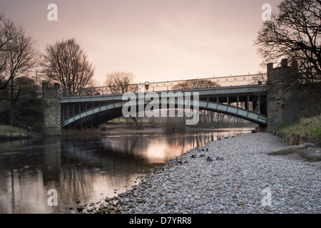 Riverside view of road bridge arching over River Wharfe at Ilkley, orange sunset glow of sky reflected in flowing water - West Yorkshire, England, UK. Stock Photo