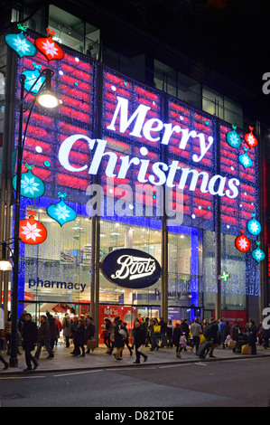 Night street scene shoppers outside shop windows Boots chemist & pharmacy store Oxford Street big Merry Christmas sign in lights London England UK Stock Photo