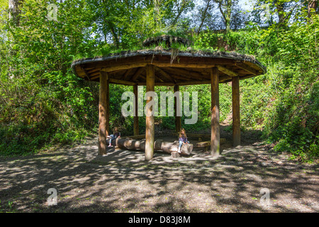 2 girls sitting under a wooden shelter with a grass roof and chimney, situated in woodland. Stock Photo