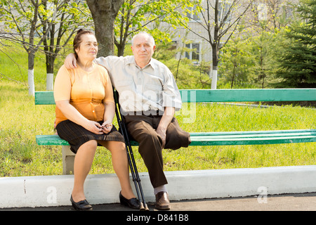 Elderly couple sitting close together on a park bench enjoying the sunshine - the husband has one leg amputated and is using crutches. Stock Photo