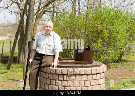 One legged man sitting on an old well in a rural park looking at the camera holding his crutches in one hand Stock Photo