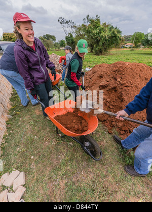 A woman waits with wheelbarrow as it is loaded with fill dirt & several other women shovel on a Habitat for Humanity build. Stock Photo