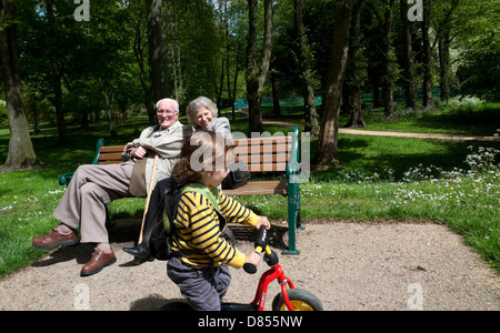 Elderly couple people grandparents sitting on bench enjoying watching a little boy child riding on his bike in Bute Park Cardiff Wales UK KATHY DEWITT Stock Photo