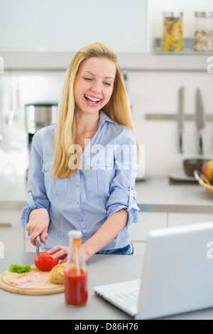 Happy teenager girl making sandwich in kitchen and looking in laptop Stock Photo
